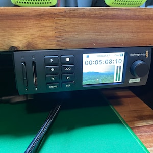 May include: Blackmagicdesign video recorder with a black and silver design. The screen displays a timer reading "00:05:08:10" and a landscape scene. The recorder has a jog wheel, buttons for play, stop, rewind, fast forward, menu, and set. There are two SD card slots labeled "1" and "2".