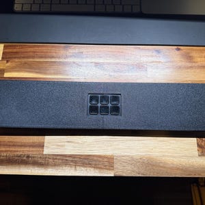 以下が含まれることがあります： 中央に6つの正方形のボタンが付いた、テクスチャ加工された表面の長方形の黒いパネル。 パネルの両端には取り付け穴があります。 これは電子部品である可能性があります。