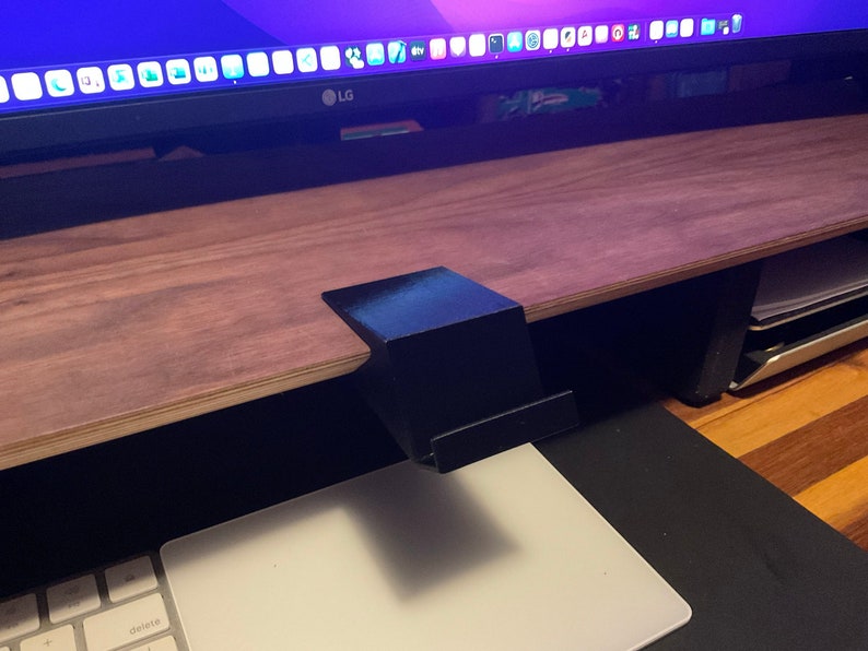 May include: A black plastic monitor stand with a flat top surface. The stand is attached to a wooden desk.