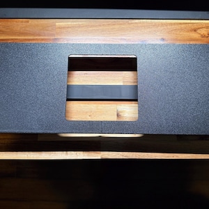 May include: A black metal panel with a textured surface and a rectangular cutout. The panel has four small holes in the corners. The panel is set on a wooden surface.