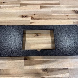 May include: A rectangular, black metal panel with a textured surface and a central rectangular cutout. The panel has rounded corners and small holes at each corner. The panel is displayed on a wooden surface.