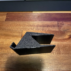 May include: A black plastic phone stand with a textured surface. The stand is designed to hold a phone upright.