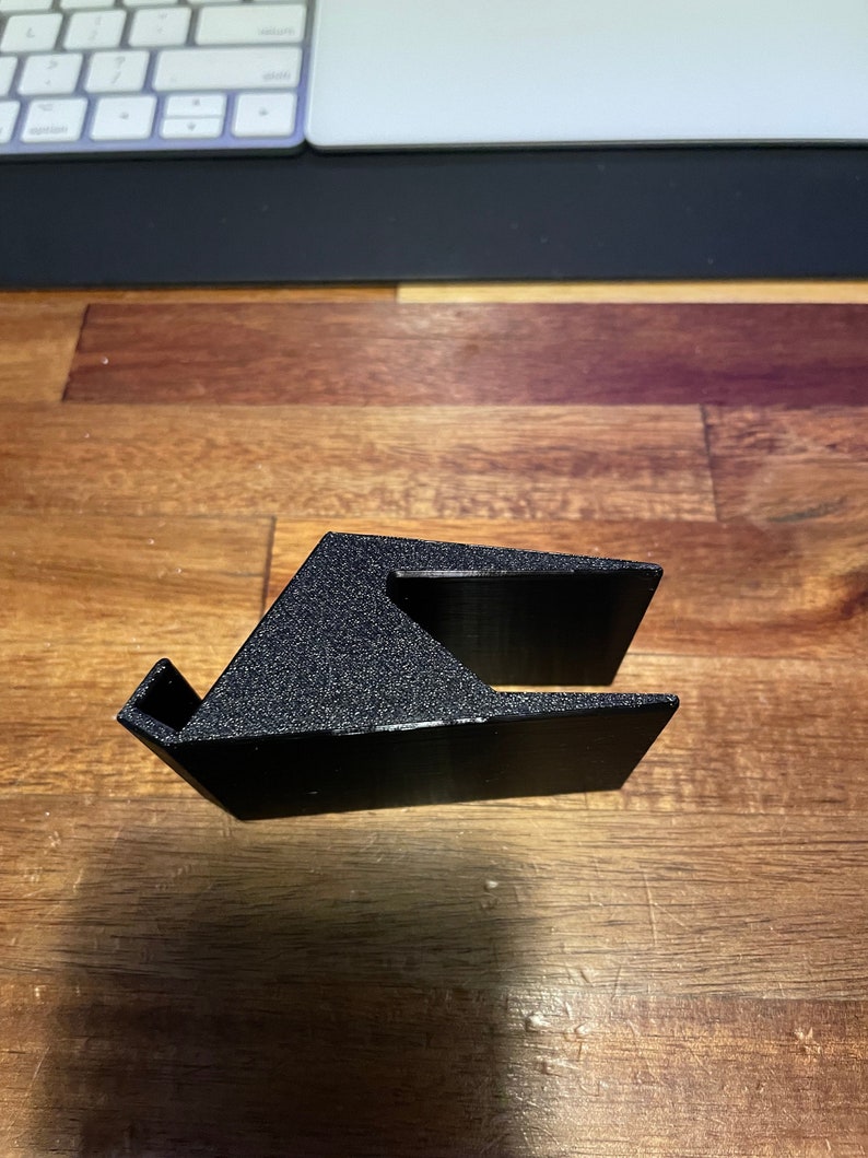 May include: A black plastic phone stand with a textured surface. The stand is designed to hold a phone upright.