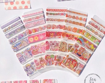 Puede incluir: Una colección de cintas washi coloridas con lindos personajes de dibujos animados y patrones. Las cintas se muestran sobre un fondo blanco y están organizadas en filas.