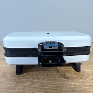 May include: A white Vitantonio Belgian Waffle maker with a black handle and black base feet. The front of the appliance features a silver and blue label with the text Vitantonio Belgian Waffle. The unit is rectangular with rounded corners.