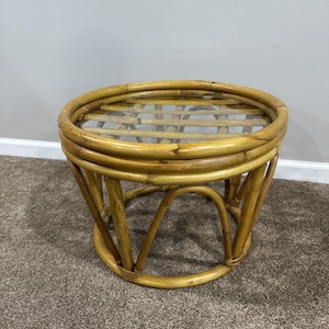May include: A round, natural-toned rattan side table with a clear glass top. The table has a woven design and curved legs, creating a vintage aesthetic. The table is approximately 18 inches in diameter and 18 inches tall.