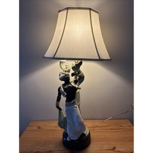 May include: A vintage table lamp featuring a black-skinned figure holding a stylized serpent, with a cream-colored lampshade. The base is dark green, and the figure is adorned with gold accents. The lamp is on a wooden surface.