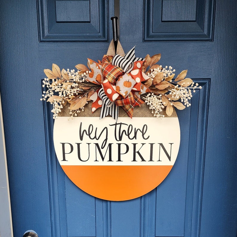 May include: A round wooden door hanger with a white background and orange bottom half. The hanger has the text 'hey there PUMPKIN' in black lettering. The hanger is decorated with a brown, white, and orange bow with a plaid pattern and small white pumpkins. The hanger is hanging on a blue door.