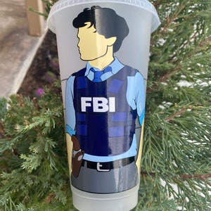 May include: A clear plastic tumbler with a design of a cartoon character wearing a blue shirt, a blue vest with the word "FBI" on it, and a gun in a holster. The character has black hair and a yellow face.