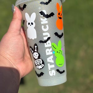 May include: A clear Starbucks cold cup with a glittery finish, decorated with Halloween-themed bunny and bat decals. The cup features a white bunny, a skeleton bunny, an orange pumpkin bunny, and a green bunny. Black bat silhouettes are scattered around the cup.