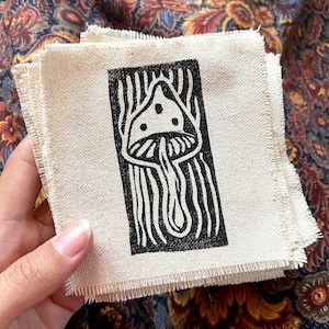 May include: A black and white illustration of a mushroom printed on a white fabric patch. The mushroom has a rounded cap with a stem and is surrounded by a black and white striped pattern.