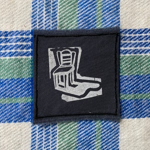 May include: A black fabric patch with a white silhouette of a chair. The chair has a back and four legs. The patch has a white border.