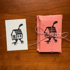 May include: A black and white illustration of a house with legs printed on a white card and a pink card. The pink card is wrapped with white twine.