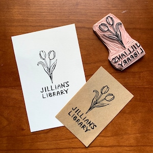 May include: A black and white tulip design stamp with the text "Jillian's Library" is next to two cards with the same design. One card is white, the other is brown. The stamp is pink.