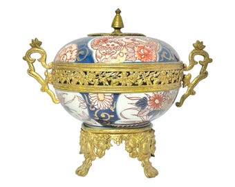 May include: A decorative ceramic bowl with a lid, featuring a blue and white floral design with red accents. The bowl has a gold-colored metal filigree band and handles, and sits on a matching ornate stand. The lid is topped with a gold finial.