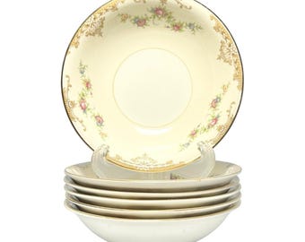 May include: A set of six cream-colored vintage soup bowls. Each bowl features a gold rim and floral and gold filigree detailing. The bowls are stacked and displayed on a white background, showcasing their elegant design.