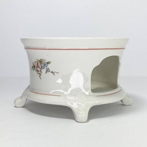 May include: White ceramic tea warmer with a floral design and three legs. The warmer has a cut-out section for a tea light.