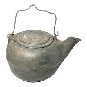 May include: Antique metal teapot with a curved handle and a spout. The lid features a starburst design. The teapot has a dark, aged appearance, suggesting it is vintage. The handle is made of wire. The teapot is likely used for brewing tea or other hot beverages.