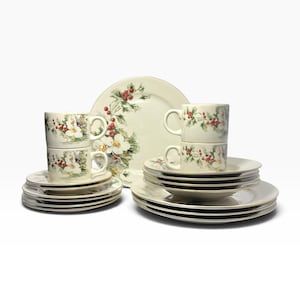 May include: A dinnerware set featuring off-white ceramic plates, saucers, and mugs decorated with a festive pattern of white flowers, green holly leaves, and red berries. The pieces have gold-toned rims and are arranged in stacks on a white surface.