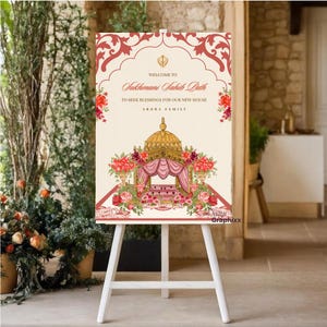 May include: A rectangular welcome sign on a white easel featuring a cream background with red floral borders. The text reads: Welcome to Sukhmani Sahib Path to seek blessings for our new house Arora Family. A gold and pink illustration is centered.