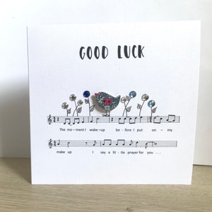 May include: A white card with the words "GOOD LUCK" printed in black. A small bird with a floral pattern is perched on a musical staff with the lyrics "The moment I wake-up before I put on my make up I say a little prayer for you..." printed below.