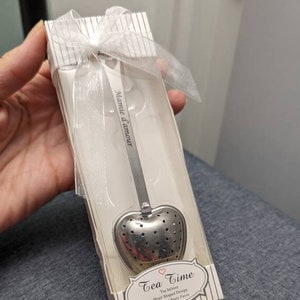 Può includere: Una scatola bianca con un infusore per tè a forma di cuore argentato. La scatola è decorata con un nastro bianco e le parole "Tea Time" e "Mamie d'amour" stampate su di essa.