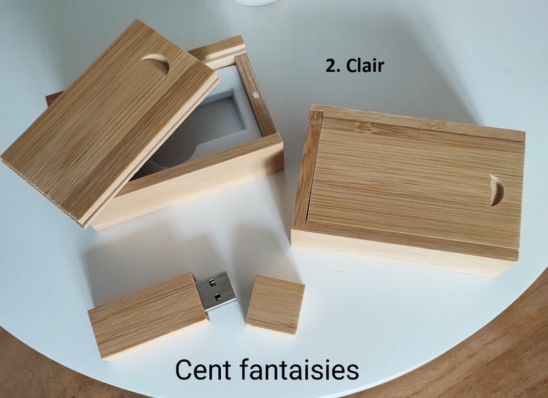 Op de afbeelding: Twee houten dozen met deksels, een open en een gesloten, en een houten USB-stick met een metalen connector. De dozen zijn gemaakt van lichtbruin hout met een natuurlijke nerf. De USB-stick is ook gemaakt van lichtbruin hout. De tekst "Cent fantaisies" is afgedrukt op het witte oppervlak onder de items.