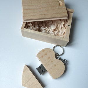 Op de afbeelding: Een houten hartvormige USB-stick met een sleutelhanger. De stick is open, waardoor de USB-connector zichtbaar is. De stick zit in een kleine houten doos met een deksel.