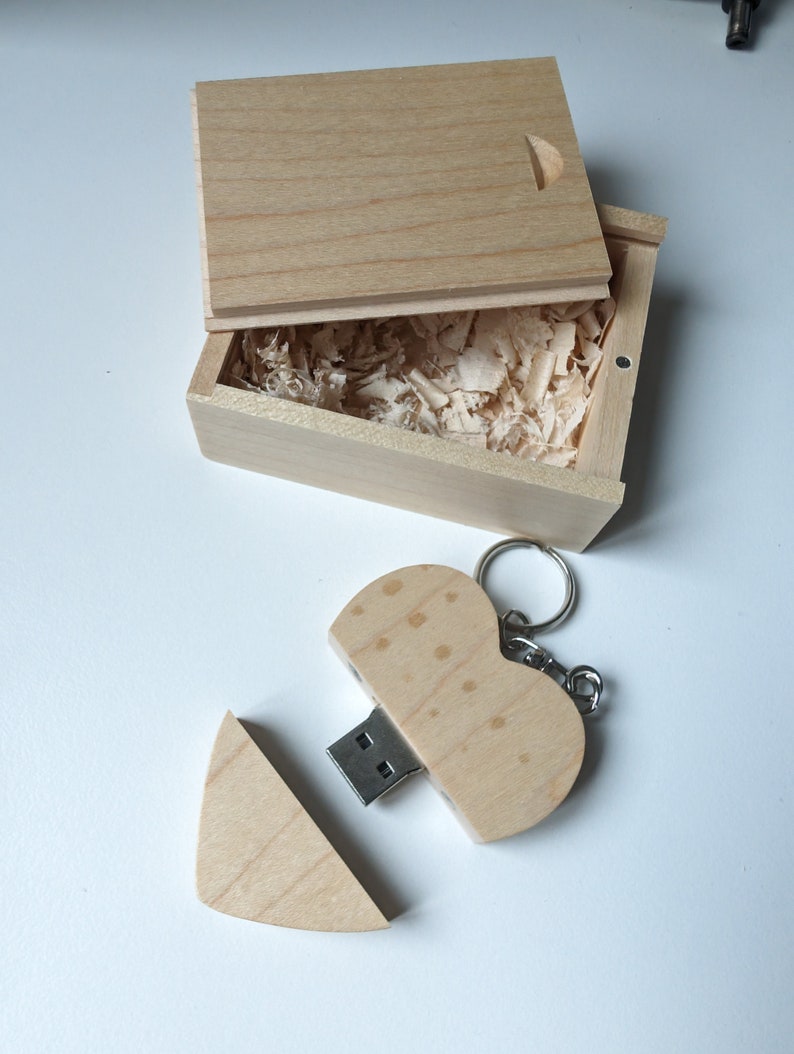 Op de afbeelding: Een houten hartvormige USB-stick met een sleutelhanger. De stick is open, waardoor de USB-connector zichtbaar is. De stick zit in een kleine houten doos met een deksel.