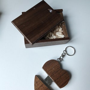 Op de afbeelding: Een houten hartvormige USB-stick met een sleutelhanger. De stick is open, waardoor een USB-connector zichtbaar is. De stick zit in een kleine houten doos met een deksel.