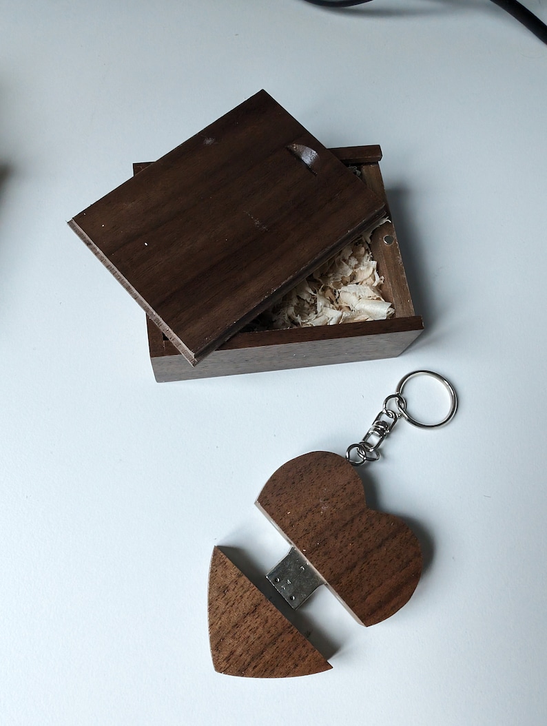 Op de afbeelding: Een houten hartvormige USB-stick met een sleutelhanger. De stick is open, waardoor een USB-connector zichtbaar is. De stick zit in een kleine houten doos met een deksel.