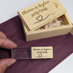 Op de afbeelding: Een houten doos met een deksel gegraveerd met "Christian & Guyliane 21/07/2023" en een hartontwerp. De doos is open en bevat een kleine, transparante USB-stick van acryl met dezelfde gravure.
