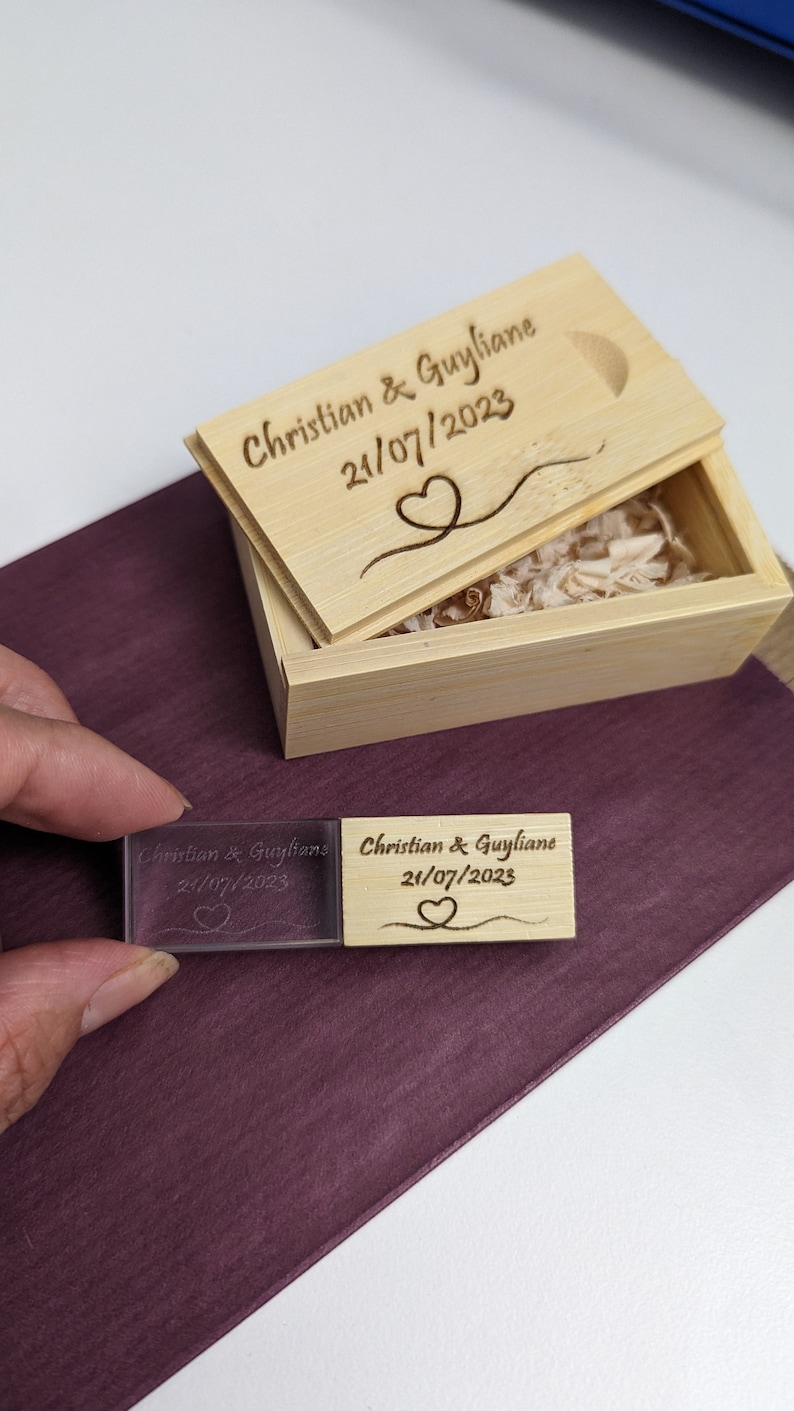 Op de afbeelding: Een houten doos met een deksel gegraveerd met "Christian & Guyliane 21/07/2023" en een hartontwerp. De doos is open en bevat een kleine, transparante USB-stick van acryl met dezelfde gravure.