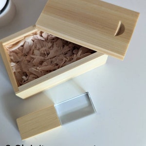 Op de afbeelding: Een houten doos met een deksel, gevuld met houtsnippers, en een transparante glazen USB-stick met een houten basis. De tekst "3.Clair/transparent" is zichtbaar onder de doos.