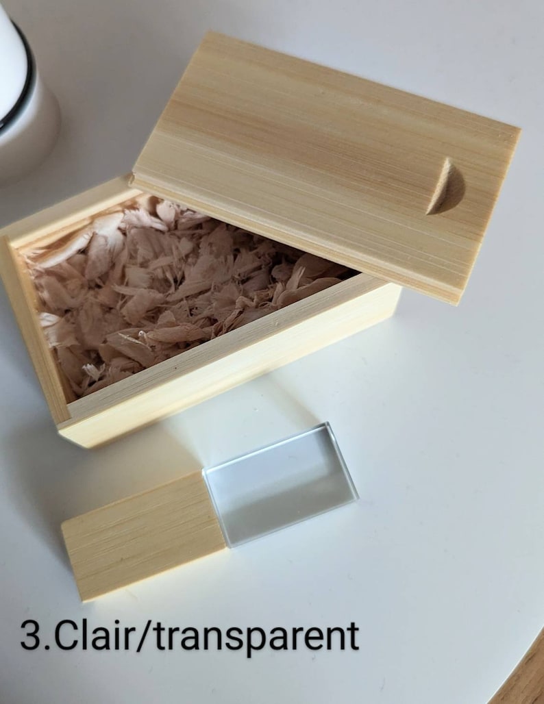 Op de afbeelding: Een houten doos met een deksel, gevuld met houtsnippers, en een transparante glazen USB-stick met een houten basis. De tekst "3.Clair/transparent" is zichtbaar onder de doos.