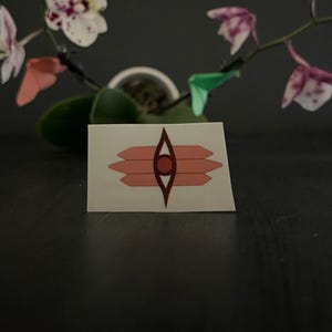 May include: A rectangular sticker featuring a geometric design. The design includes a central eye-like shape in dark red, surrounded by three horizontal, light red, arrow-like shapes on each side. The sticker is on a dark surface, with a blurred orchid in the background.