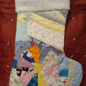 May include: A patchwork Christmas stocking with a white faux fur cuff. The stocking is made of various colorful fabrics, including blue, yellow, pink, and green. The stocking features a large, colorful flower design in the center.