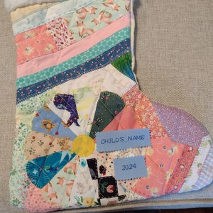 May include: A patchwork Christmas stocking with a white faux fur cuff. The stocking is decorated with a variety of colorful fabrics and patterns, including floral prints, geometric designs, and animal motifs. The stocking has a blue tag with the text "CHILD'S NAME" and a blue tag with the text "2024".