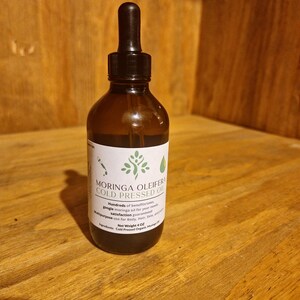 May include: A dark brown glass bottle with a dropper top filled with a dark liquid. The bottle has a white label with the text "MORINGA OLEIFERA COLD PRESSED OIL" and information about the product. The bottle is sitting on a wooden surface.