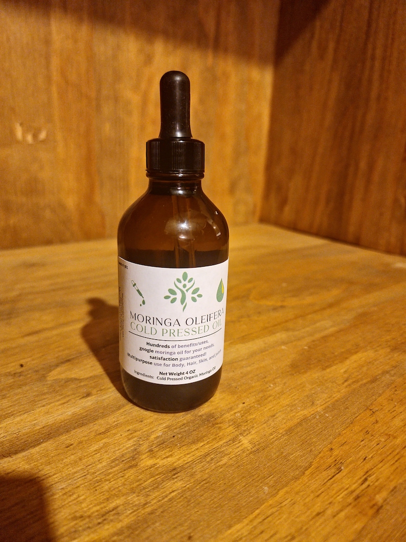 May include: A dark brown glass bottle with a dropper top filled with a dark liquid. The bottle has a white label with the text "MORINGA OLEIFERA COLD PRESSED OIL" and information about the product. The bottle is sitting on a wooden surface.
