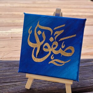 May include: A small blue canvas painting with gold Arabic calligraphy on a wooden easel.