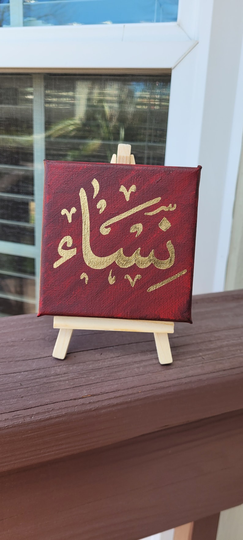 May include: A small canvas painting on a wooden easel with a gold Arabic calligraphy design on a red background.