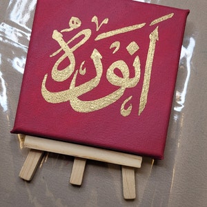 May include: A red canvas with a gold Arabic calligraphy design on an easel. The calligraphy says "Allah" in Arabic.