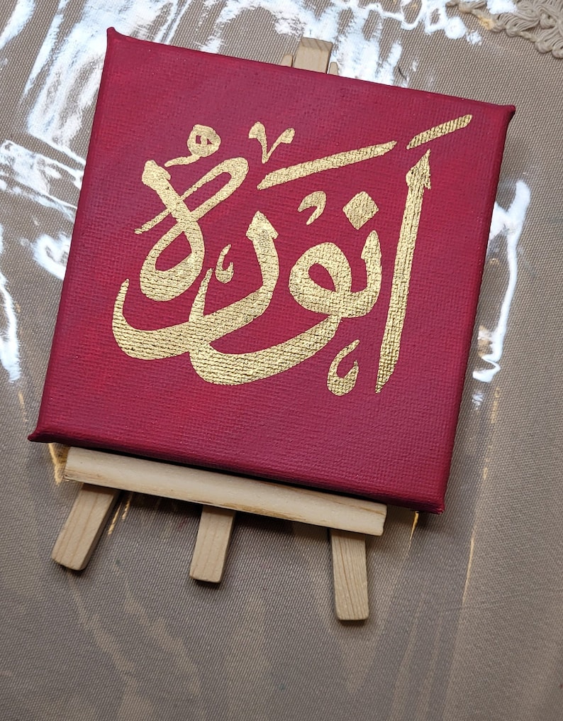 May include: A red canvas with a gold Arabic calligraphy design on an easel. The calligraphy says "Allah" in Arabic.