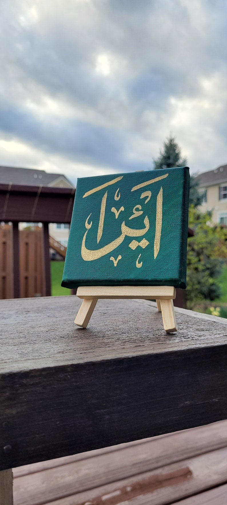 May include: A small green canvas with gold Arabic calligraphy on an easel. The calligraphy reads "أَقْبَلَ".