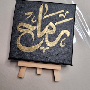 May include: A small black canvas with a gold Arabic calligraphy design on an easel.