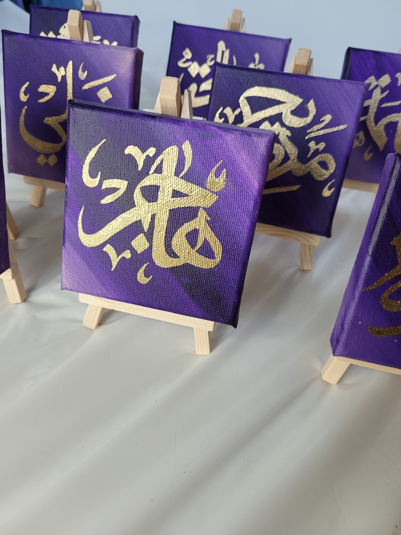 May include: A set of small purple canvas paintings with gold calligraphy on easels. The paintings feature different Arabic words.