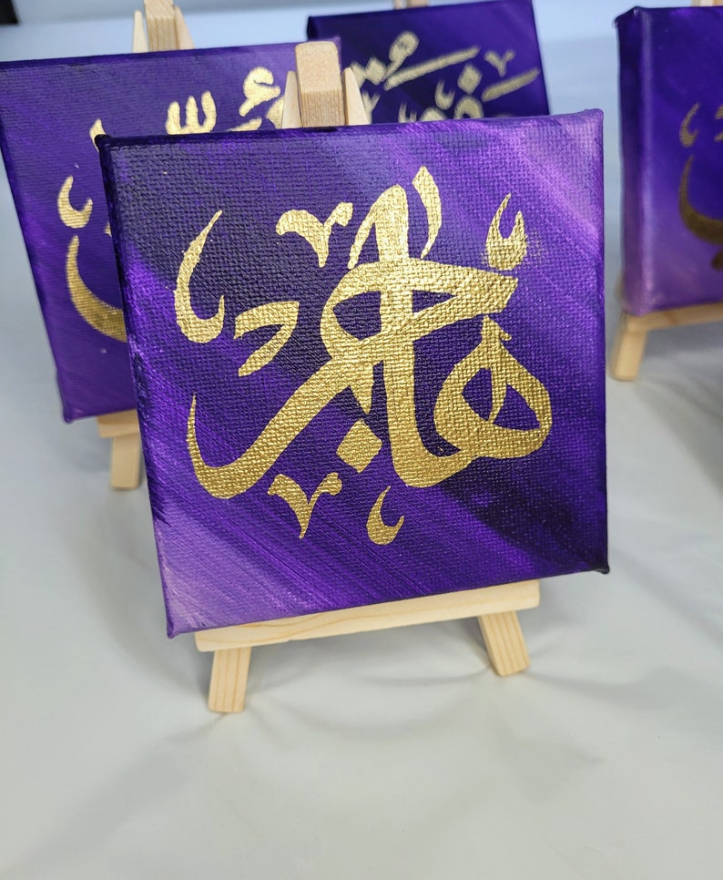 May include: A small purple canvas painting with gold Arabic calligraphy on a wooden easel. The calligraphy is in a decorative style and the background is a gradient of purple shades.