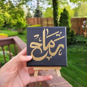 May include: A small black canvas with gold calligraphy on an easel. The calligraphy says "شكرًا" which is Arabic for "thank you".