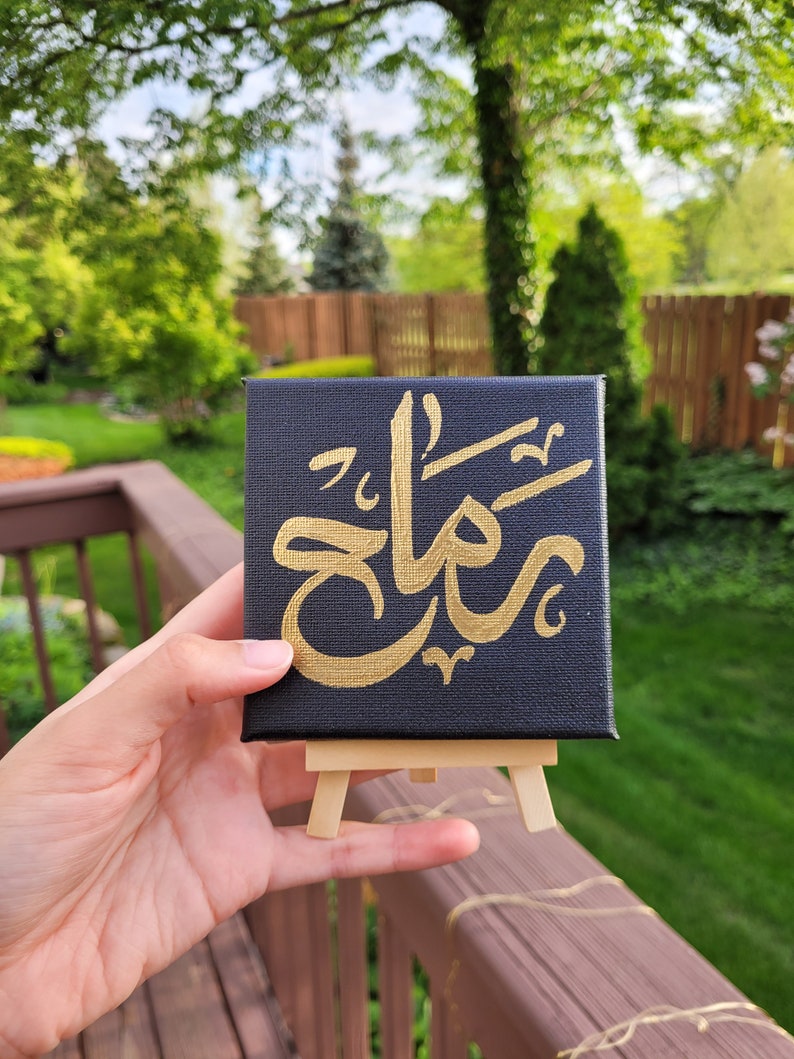 May include: A small black canvas with gold calligraphy on an easel. The calligraphy says "شكرًا" which is Arabic for "thank you".